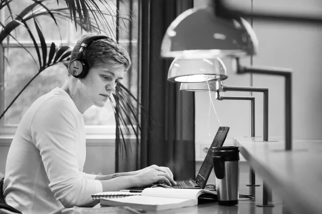 A student with headphones is working on his laptop under light fixtures. Photographer Johan Bävman.