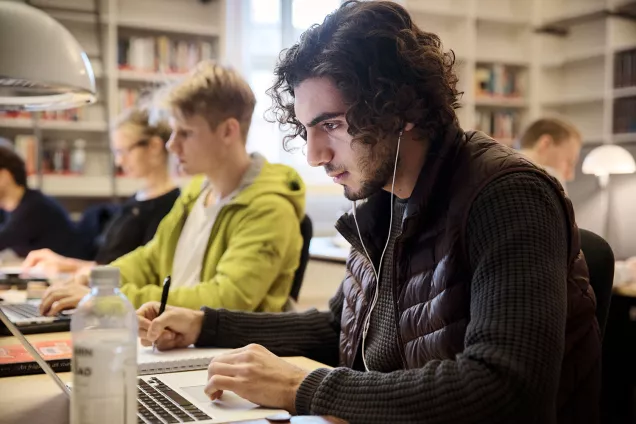 Students working on their laptops along a table. White bookshelves in the background. Photographer Johan Bävman.