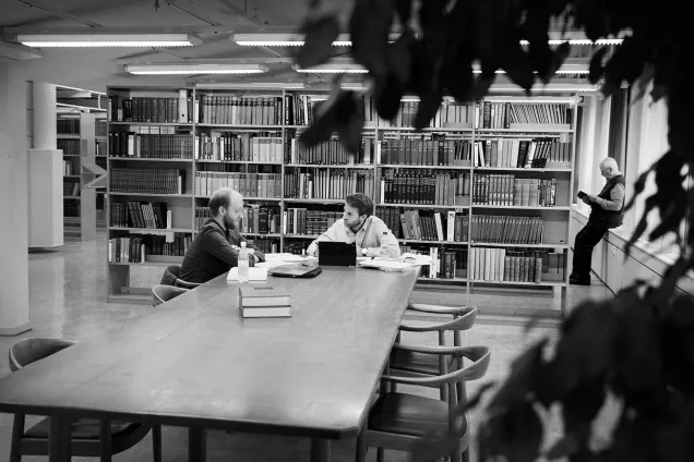 Two students work at a table with bookshelves behind them. Photographer Johan Bävman.