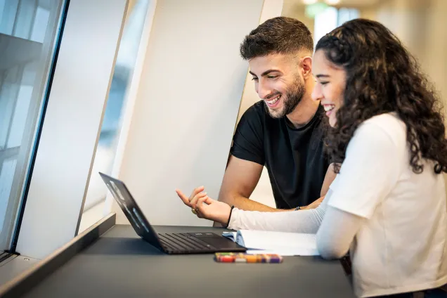 Two students are standing beside a laptop on a standing table, discussing. Photographer Johan Persson.