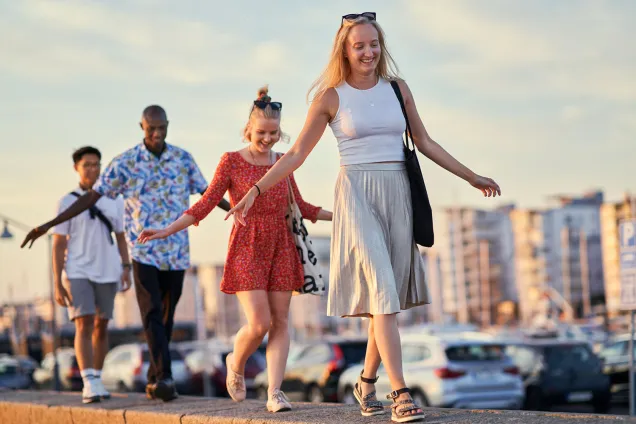 Four students are walking in a line on a stone wall next to parked cars. Photographer Johan Persson.