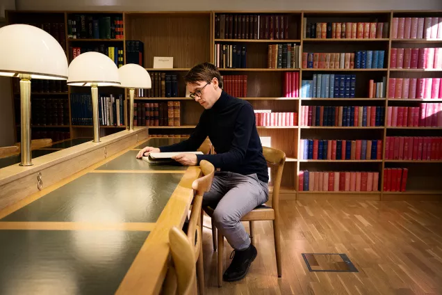 En forskare studerar dokument under opal bordslampa vid skrivbord med grönt skinn. I bakgrunden finns bokhyllor. Fotograf Johan Bävman.