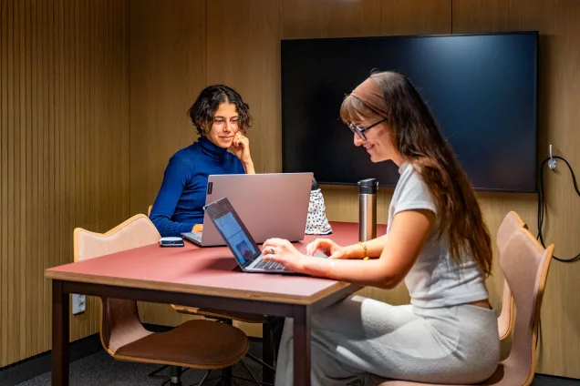 Två glada studenter arbetar vid sina laptops i ett rum med fyra stolar och skärm på väggen..